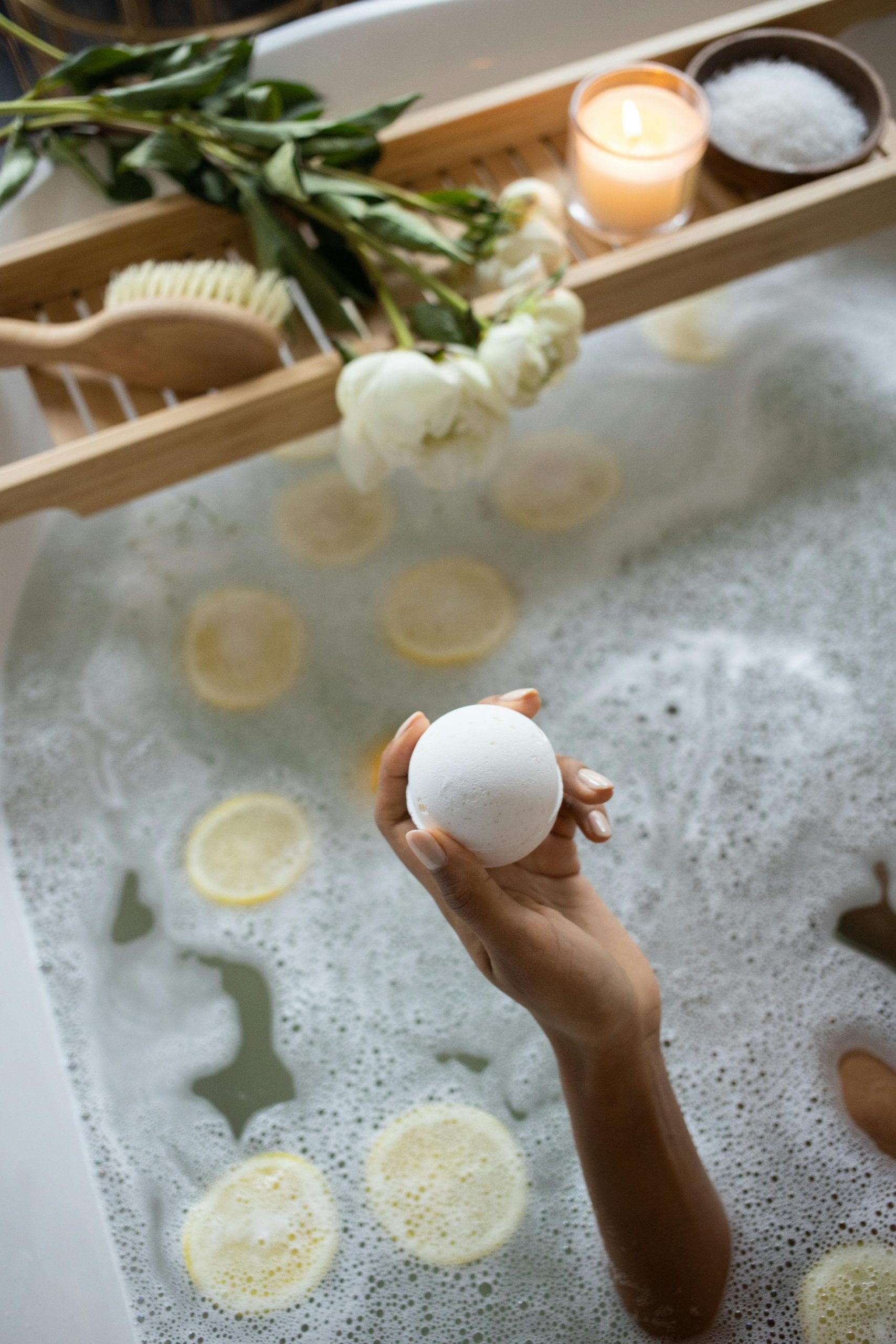 From above of unrecognizable female sitting in bath with lemon slices on water surface near wooden tray with salt and burning candle decorated with flowers during daily routine